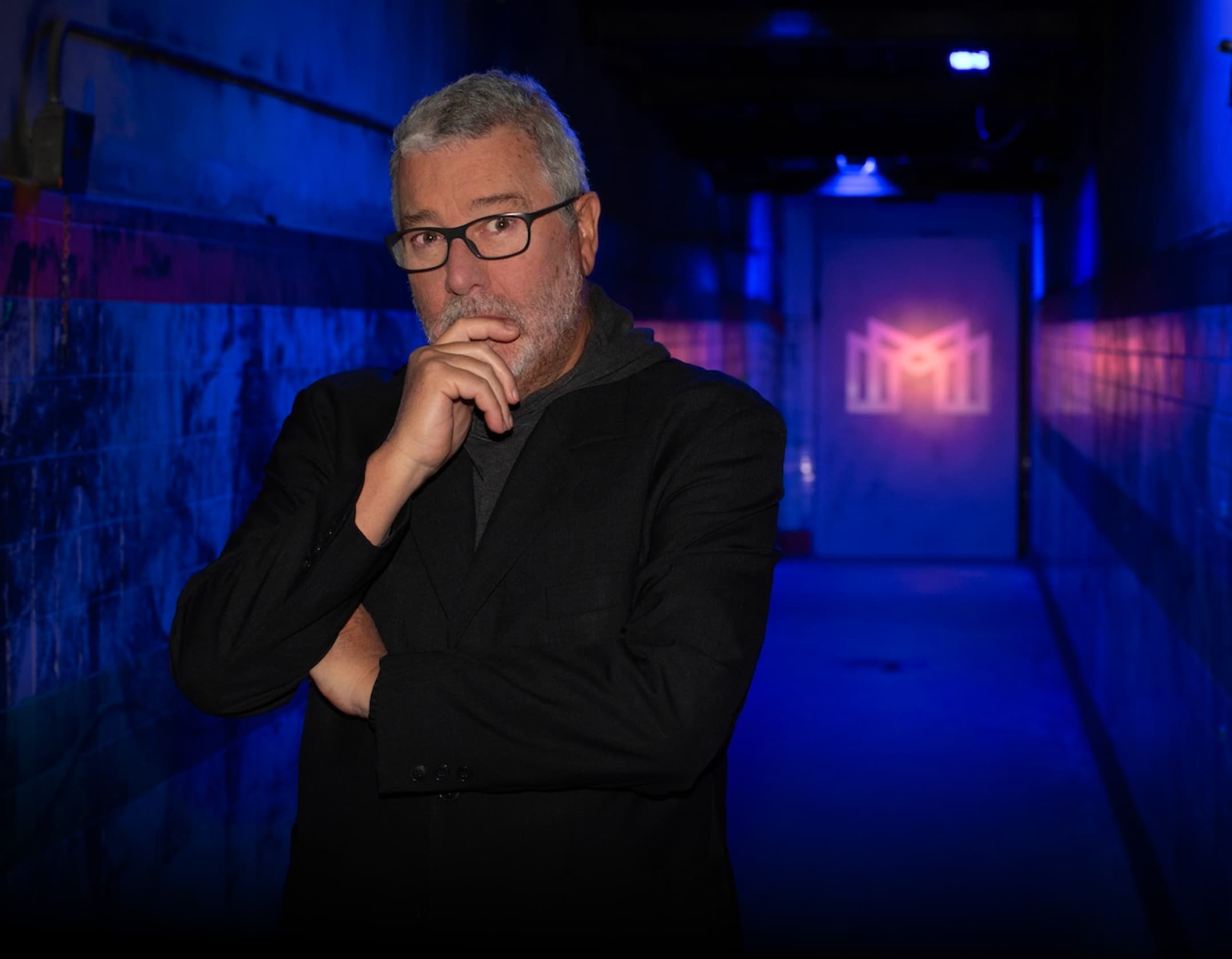 The image features Philippe Starck standing in a pensive pose, against the backdrop of a luxurious, dimly lit corridor bathed in deep, immersive blue lighting. The scene is enriched by the soft glow of the iconic Mortlach 'M' monogram elegantly projected onto a sleek door in the distance, creating an aura of exclusivity and modern elegance.