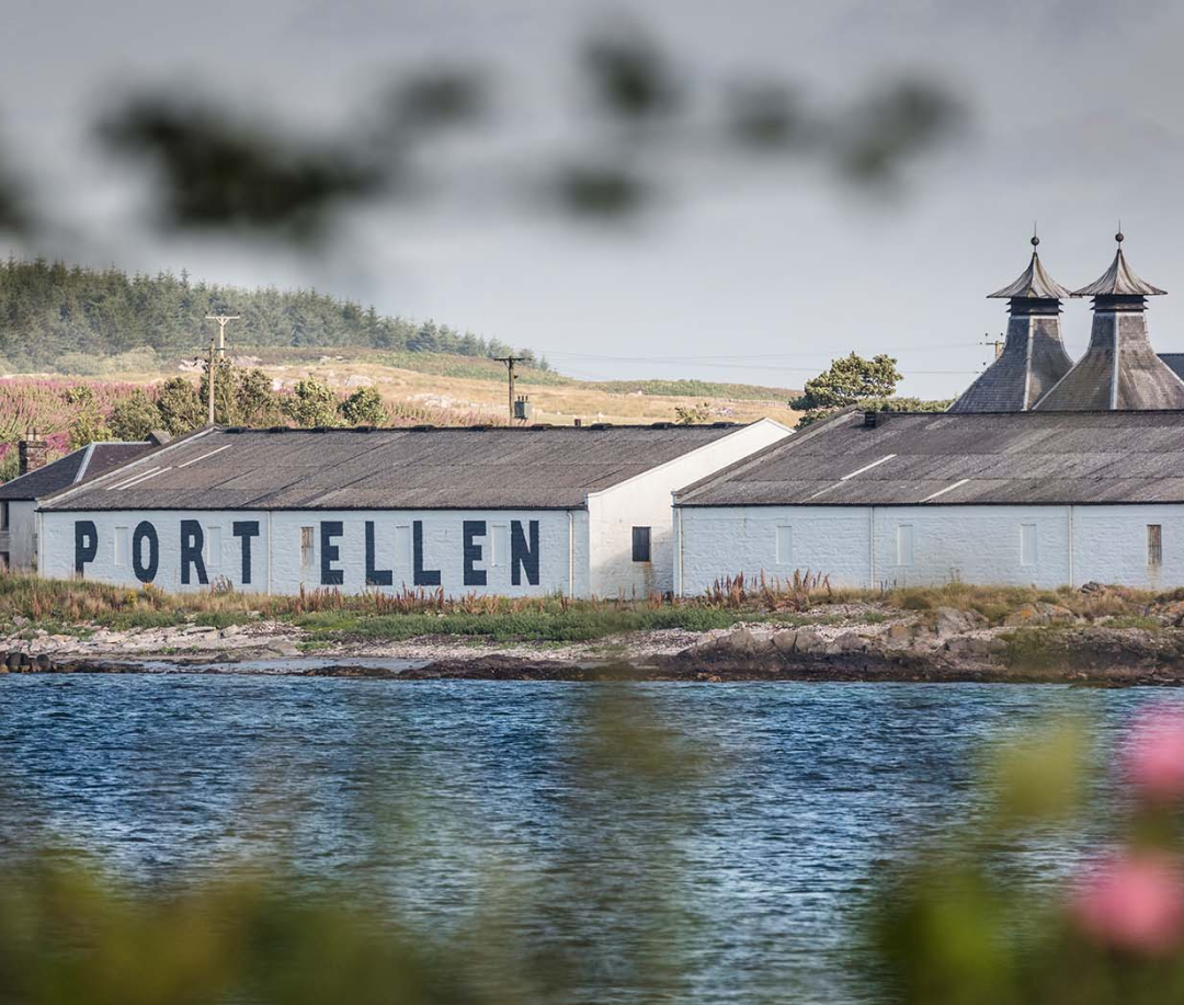 A scenic view of the historic Port Ellen distillery, with white-washed buildings and dark slate rooftops along a tranquil shoreline. The name 'PORT ELLEN' is boldly painted on one of the buildings. In the background, rolling green hills and trees enhance the peaceful landscape, while the calm water and soft greenery in the foreground add to the rustic charm.